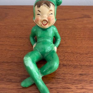 Könnte beinhalten: Eine Keramikfigur eines lachenden Elfen, der einen leuchtend grünen Anzug und eine passende spitze Mütze trägt. Der Elf hat braune Haare, rosige Wangen und einen offenen Mund. Die Figur ist in einer sitzenden Position mit gekreuzten Beinen dargestellt.