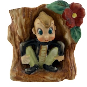 Può includere: Statuina vintage in ceramica di un ragazzo seduto, incastonato in un tronco d'albero. Il ragazzo indossa una giacca nera, pantaloni verdi e scarpe nere. Un fiore rosso con foglie verdi adorna il lato. La statuina ha una combinazione di colori marrone chiaro e beige.