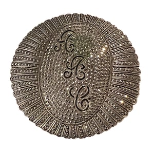 May include: A silver oval brooch with a raised design of the letters "A C C" in a stylized font. The brooch is covered in small, round, silver-toned stones.