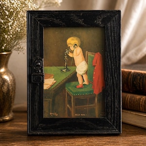 May include: A framed vintage print shows a baby using a telephone. The artwork features a baby in a nappy standing on a green chair, with a red coat hanging nearby. The frame is black and the text "HELLO PAPA!" is visible.