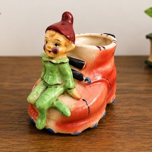 May include: A ceramic planter shaped like a shoe with a small elf figure sitting on the front. The elf wears a red hat and a green outfit. The shoe is primarily orange with black laces and blue accents along the base. The interior is cream colored.