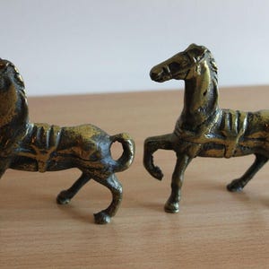 May include: Two small, antique-style brass horse figurines standing in profile. Each horse features a textured, weathered metallic finish, a molded saddle, and a raised front leg. The figures are cast in a solid, dark gold-toned metal alloy.