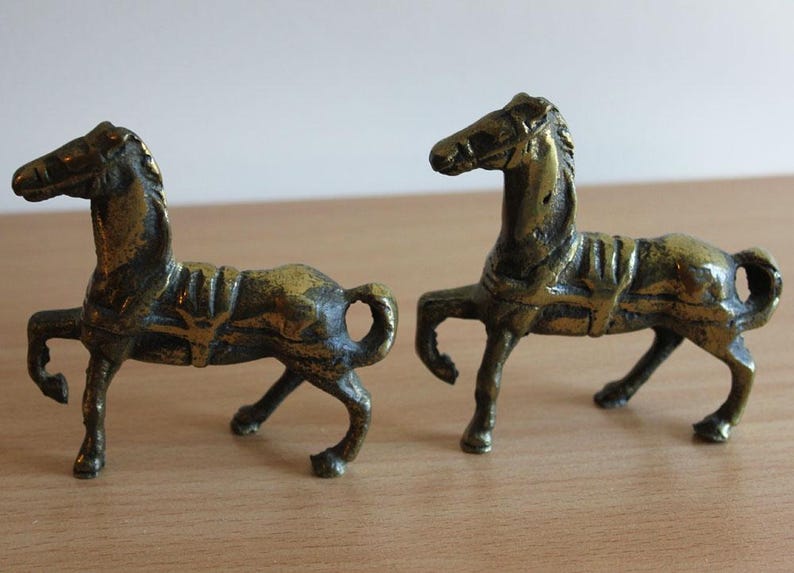 May include: Two small, antique-style brass horse figurines standing in profile. Each horse features a textured, weathered metallic finish, a molded saddle, and a raised front leg. The figures are cast in a solid, dark gold-toned metal alloy.