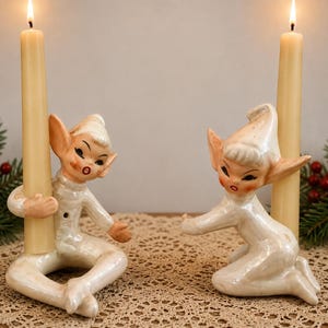 Op de afbeelding: Twee keramische elfenkaarsenhouders met een glanzende witte afwerking, elk met puntige oren en een muts. De elfen zitten in een zittende houding en houden lange, slanke, lichtbeige taps toelopende kaarsen vast die branden met een kleine vlam.
