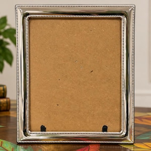 May include: A rectangular picture frame with a polished silver-tone metallic finish and a beaded border detail. The frame features a plain, light brown cardboard backing visible through the center, with two small black clips at the bottom edge.
