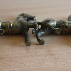 May include: Two small, metallic, bronze-colored horse figurines shown from a side profile. Each horse is depicted in a standing position with detailed musculature, a saddle, and a textured finish, capturing a realistic equine form.