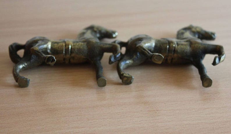 May include: Two small, metallic, bronze-colored horse figurines shown from a side profile. Each horse is depicted in a standing position with detailed musculature, a saddle, and a textured finish, capturing a realistic equine form.