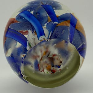 Peut inclure: Un presse-papier en verre transparent avec des motifs internes color&eacute;s. Le presse-papier pr&eacute;sente des rubans et des tourbillons bleus, orange et blancs, avec une spirale verte &agrave; la base. De petites bulles d'air sont visibles dans le verre.