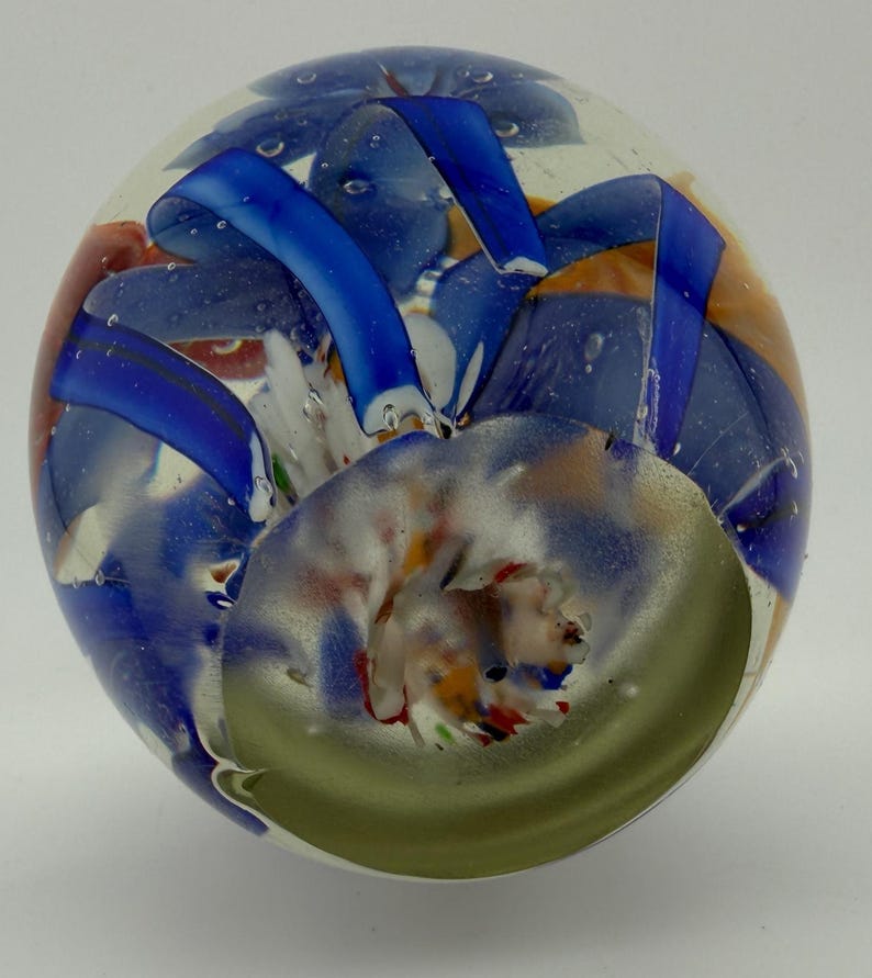 Peut inclure: Un presse-papier en verre transparent avec des motifs internes color&eacute;s. Le presse-papier pr&eacute;sente des rubans et des tourbillons bleus, orange et blancs, avec une spirale verte &agrave; la base. De petites bulles d'air sont visibles dans le verre.