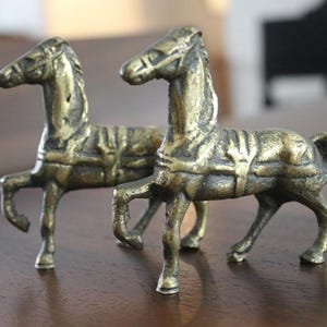May include: A pair of small, antique-style brass horse figurines. Each horse is depicted in a standing pose with one front leg raised, featuring detailed molded saddles, bridles, and manes with a weathered, dark metallic patina finish.