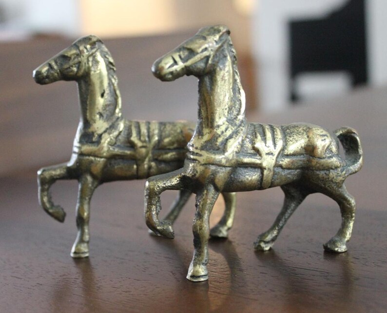 May include: A pair of small, antique-style brass horse figurines. Each horse is depicted in a standing pose with one front leg raised, featuring detailed molded saddles, bridles, and manes with a weathered, dark metallic patina finish.