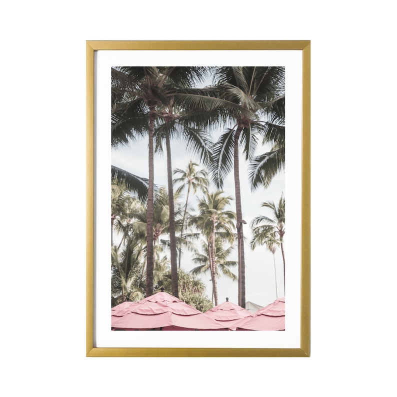 Op de afbeelding: Een ingelijste print van een tropische sc&egrave;ne met palmbomen en roze parasols. De print heeft een gouden lijst.