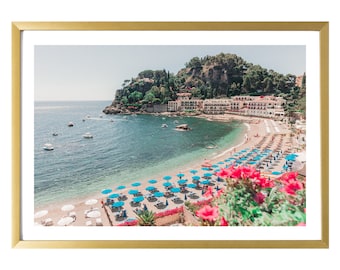 May include: A scenic view of a beach with blue umbrellas and lounge chairs, a small town in the background, and a clear blue ocean with boats in the distance.