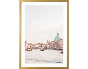 May include: A framed photograph of a canal in Venice, Italy. The photo shows a view of the Grand Canal with buildings lining the sides and a bridge in the distance. The water is calm and the sky is a light blue.
