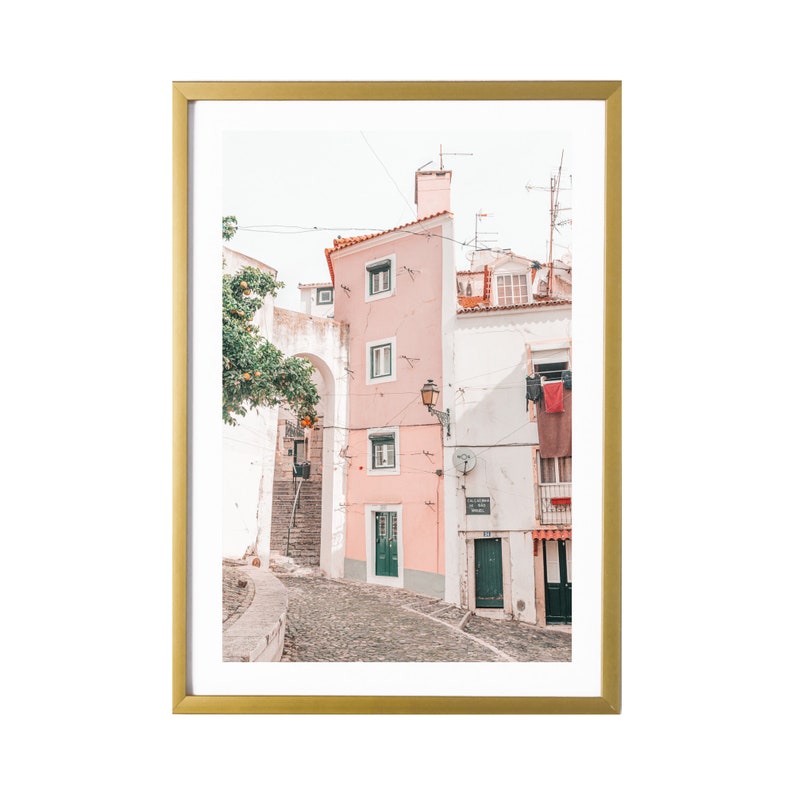 May include: A photo of a narrow, cobblestone street in a European city. The buildings are painted in shades of pink and white, with green doors and windows. There are clothes hanging on a line in the background.