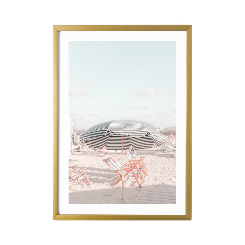 May include: A framed photograph of a beach scene with striped beach umbrellas and red and white beach chairs on a white sandy beach.