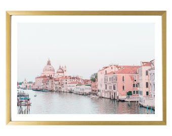 May include: A photograph of a canal in Venice, Italy. The canal is lined with pink and white buildings, and there are boats in the water. The sky is a light blue color.