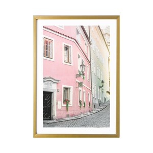 May include: A photo of a narrow, cobblestone street in a European city. The street is lined with pink and yellow buildings, some with black doors and windows. There are potted plants and streetlights on the buildings. The street curves to the right and disappears into the distance.