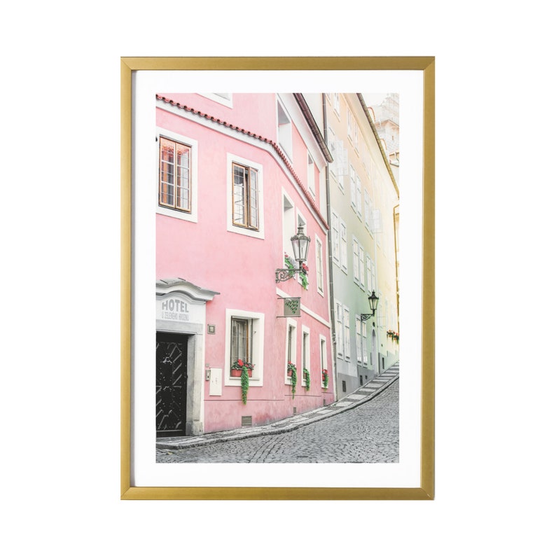 May include: A photo of a narrow, cobblestone street in a European city. The street is lined with pink and yellow buildings, some with black doors and windows. There are potted plants and streetlights on the buildings. The street curves to the right and disappears into the distance.