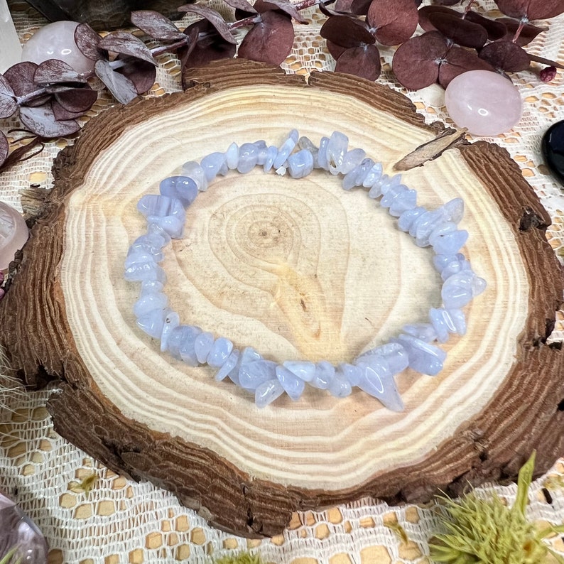 May include: A bracelet made of light blue gemstone chips, presented on a rustic wooden slice. The irregular stones create a textured appearance. The bracelet is a delicate piece of jewellery, showcasing the natural colour of the stones.