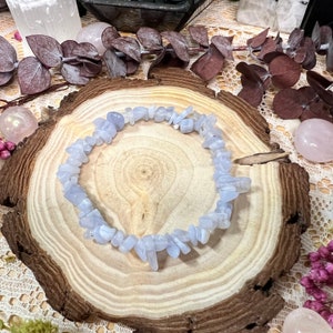 May include: A light blue chip bead bracelet. The bracelet is made of small, irregularly shaped beads. The bracelet is displayed on a wooden slice, surrounded by decorative elements including crystals and dried flowers.