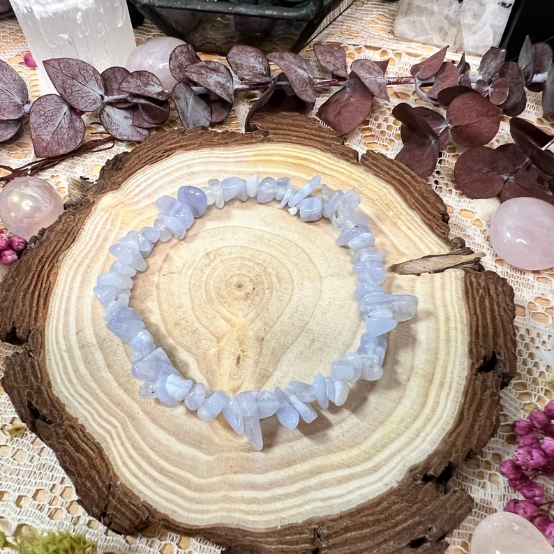 May include: A light blue chip bead bracelet. The bracelet is made of small, irregularly shaped beads. The bracelet is displayed on a wooden slice, surrounded by decorative elements including crystals and dried flowers.