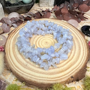 May include: A circular arrangement of pale blue tumbled gemstones, possibly blue lace agate, on a wooden slice. The gemstones are small, irregular chips, and the wood has a natural grain. The background includes dried foliage and other crystals.