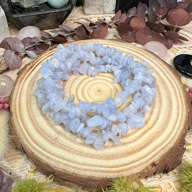 May include: A circular arrangement of pale blue tumbled gemstones, possibly blue lace agate, on a wooden slice. The gemstones are small, irregular chips, and the wood has a natural grain. The background includes dried foliage and other crystals.