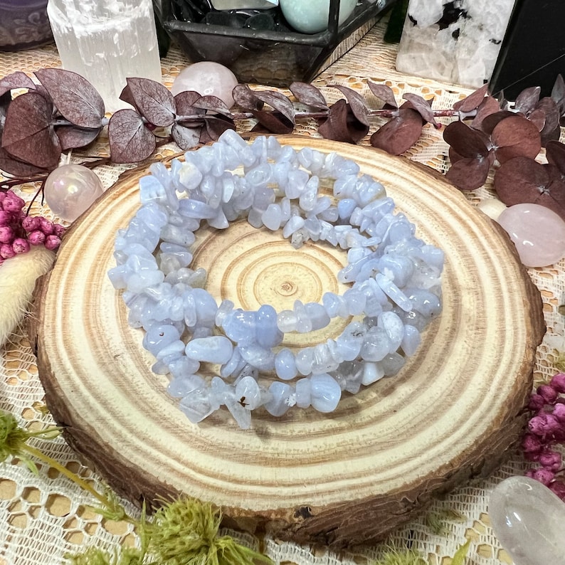 May include: A bracelet made of light blue, irregularly shaped stones. The bracelet is coiled on a round, wooden slice. The background includes dried flowers, crystals, and other decorative items.