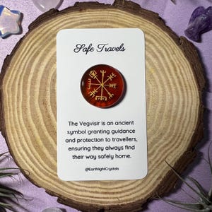 May include: A red Vegvisir symbol pin with gold detailing, presented on a white card with the words "Safe Travels." The card also has text explaining the symbol's purpose. The pin is set on a wooden slice.