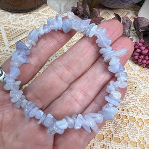 May include: A bracelet made of light blue gemstones. The bracelet is composed of small, irregularly shaped blue stones. The stones have a slightly translucent appearance. The bracelet is displayed on a person's hand.