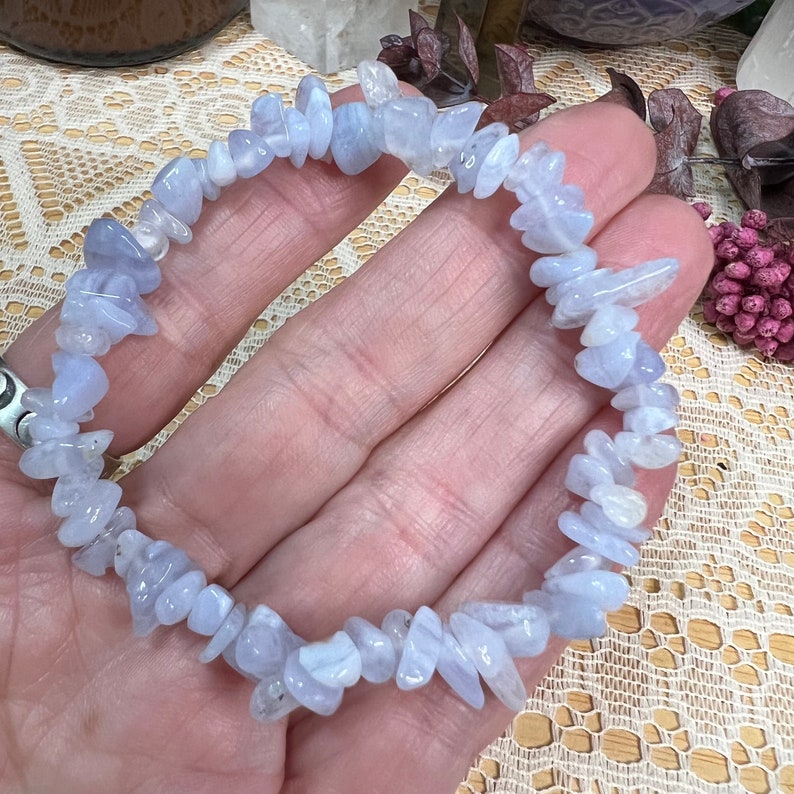 May include: A bracelet made of light blue gemstones. The bracelet is composed of small, irregularly shaped blue stones. The stones have a slightly translucent appearance. The bracelet is displayed on a person's hand.