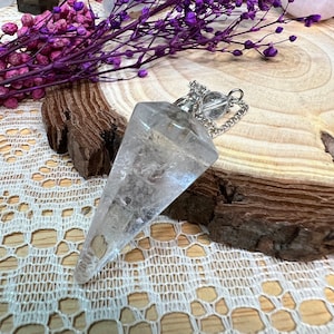 May include: A clear quartz crystal pendulum with a silver chain and a small crystal bead. The pendulum is faceted and pointed at the bottom. The background includes a wooden surface, purple dried flowers, and a piece of wood.