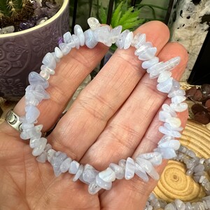 May include: A bracelet made of pale blue, irregularly shaped gemstones. The stones are strung together to form a bracelet. The gemstones have a translucent appearance. The background includes a potted plant and other decorative items.