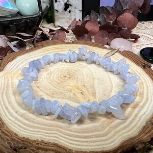 May include: A bracelet made of pale blue chip beads. The beads are small, irregularly shaped, and strung together. The bracelet has a translucent quality. It is displayed on a wooden surface with a natural grain pattern. The bracelet is approximately 18 cm in circumference.