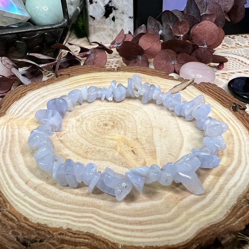 May include: A bracelet made of pale blue chip beads. The beads are small, irregularly shaped, and strung together. The bracelet has a translucent quality. It is displayed on a wooden surface with a natural grain pattern. The bracelet is approximately 18 cm in circumference.