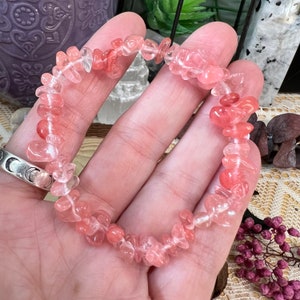 Peut inclure: Un bracelet composé de petites pierres précieuses roses et transparentes translucides et irrégulières, enfilées ensemble. Les pierres précieuses ont un aspect poli et sont disposées en cercle. Le bracelet est tenu dans une main.