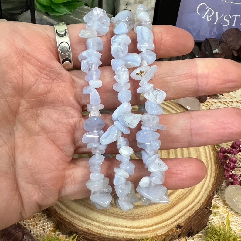 May include: Three strands of light blue gemstone chip bracelets. The bracelets are made of small, irregularly shaped stones. The bracelets are displayed on a wooden slice.