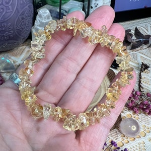 May include: A bracelet composed of small, irregularly shaped, golden-yellow citrine stones. The stones are strung together, forming a circular shape. The bracelet is held in a hand, showcasing the natural colour and texture of the gemstones.