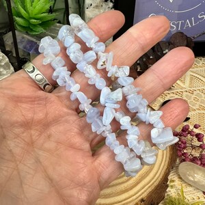 May include: A hand holds several strands of pale blue tumbled gemstones. The small, irregular chips of stone are a soft blue hue. A silver ring with a crescent moon design is visible on a finger. The gemstones are likely for crystal healing.