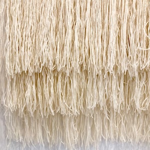 May include: A white woven wall hanging with a fringe border. The fringe is layered and has a natural, rustic look.
