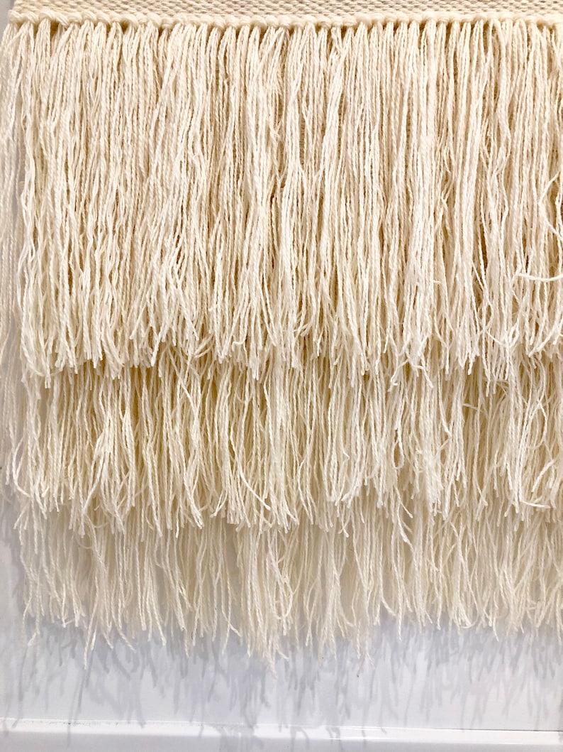 May include: A white woven wall hanging with a fringe border. The fringe is layered and has a natural, rustic look.