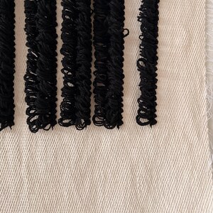 Peut inclure: Une tapisserie murale tiss&eacute;e en noir et blanc avec une bordure en franges. La tapisserie est faite de fibres naturelles et a une surface textur&eacute;e.