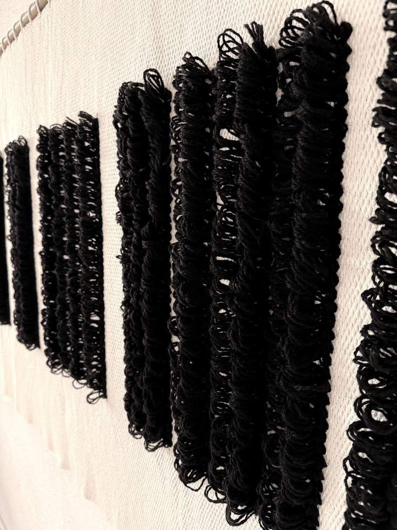 Peut inclure: Tapisserie murale noire et blanche avec une texture tiss&eacute;e. La tapisserie pr&eacute;sente une s&eacute;rie de lignes verticales noires avec un d&eacute;tail de frange.