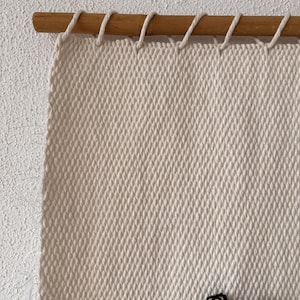 Peut inclure: Gros plan sur une tapisserie murale tiss&eacute;e beige avec une baguette en bois en haut. La tapisserie murale a un d&eacute;tail en fil noir en bas.