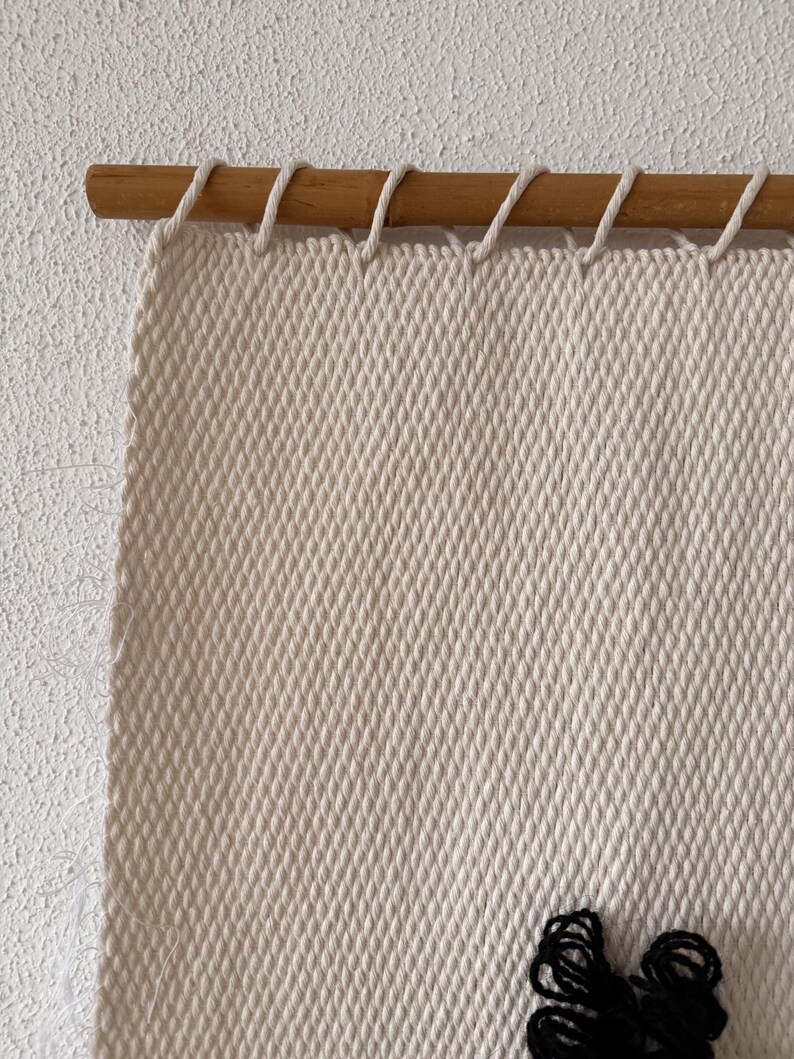 Peut inclure: Gros plan sur une tapisserie murale tiss&eacute;e beige avec une baguette en bois en haut. La tapisserie murale a un d&eacute;tail en fil noir en bas.