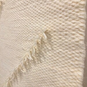 May include: Close-up of a white woven wall hanging with a textured, diagonal pattern. The edges of the hanging have a fringe.