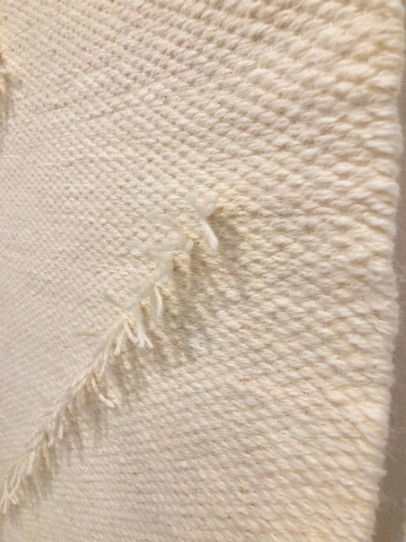 May include: Close-up of a white woven wall hanging with a textured, diagonal pattern. The edges of the hanging have a fringe.
