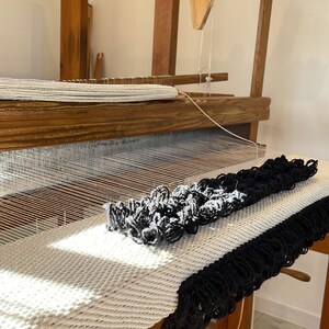 Peut inclure: Un m&eacute;tier &agrave; tisser en bois avec un tapis blanc et noir en cours de tissage. Le tapis a une frange sur le bord.