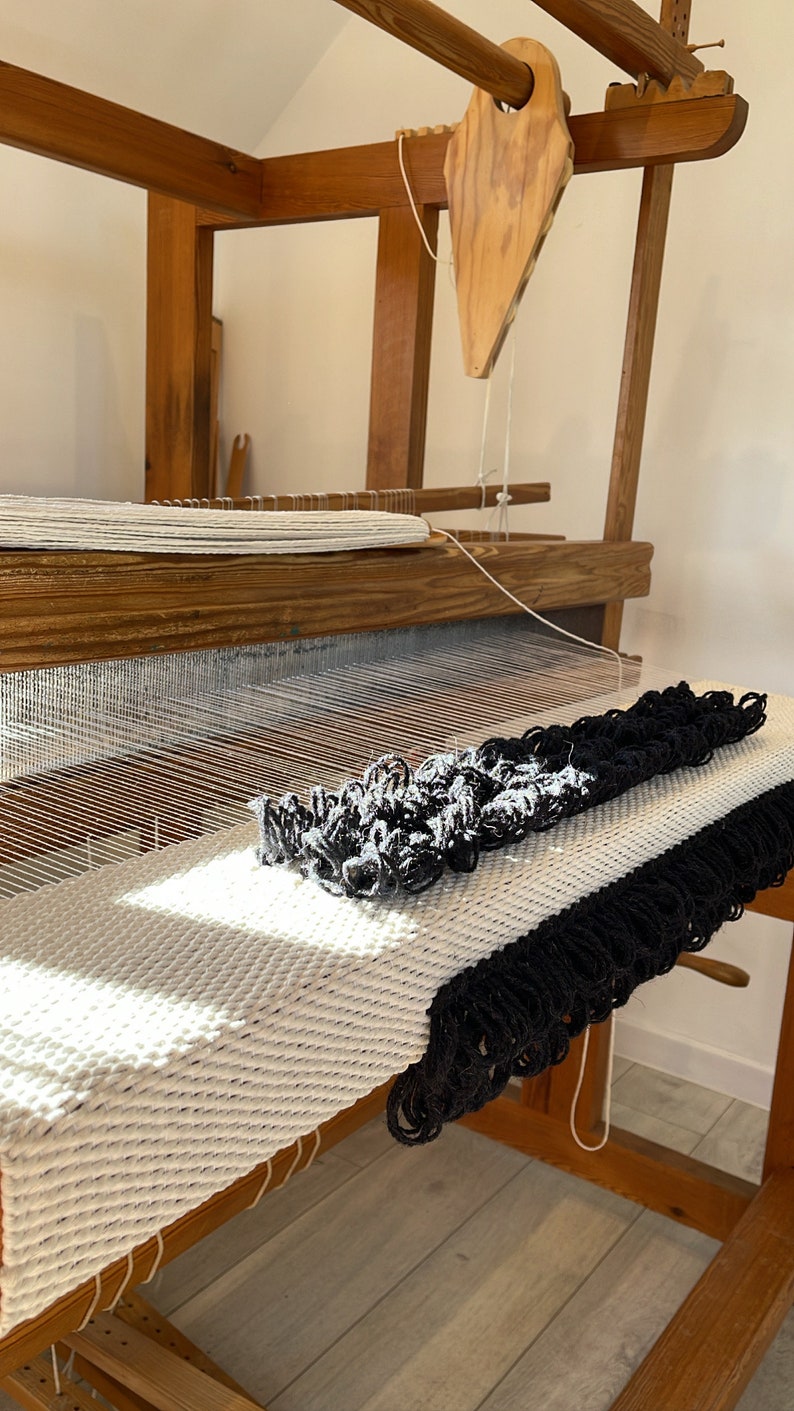 Peut inclure: Un m&eacute;tier &agrave; tisser en bois avec un tapis blanc et noir en cours de tissage. Le tapis a une frange sur le bord.